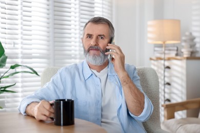 Photo of Senior man with cup of drink talking on smartphone at table indoors