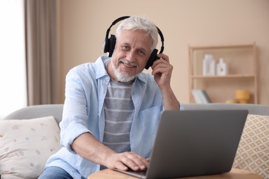 Photo of Senior man with headphones using laptop at wooden table indoors