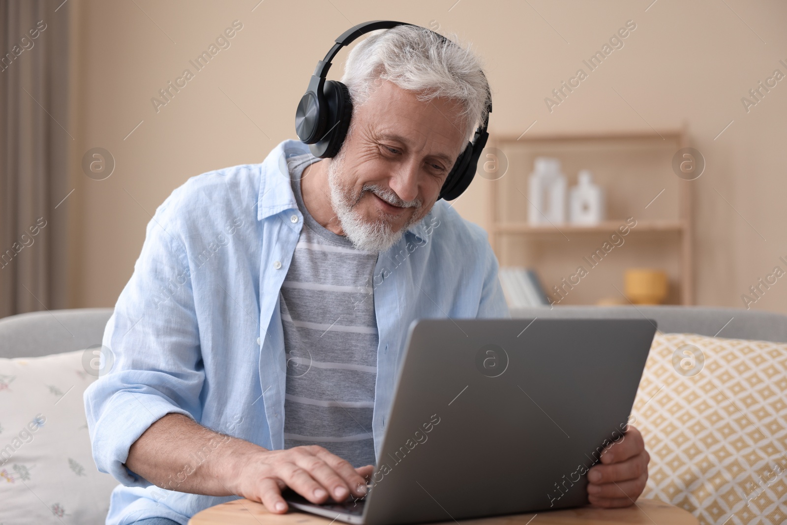 Photo of Senior man with headphones using laptop at wooden table indoors