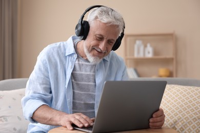 Photo of Senior man with headphones using laptop at wooden table indoors