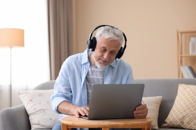 Photo of Senior man with headphones using laptop at wooden table indoors