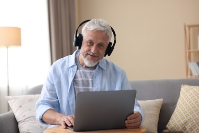 Photo of Senior man with headphones using laptop at wooden table indoors