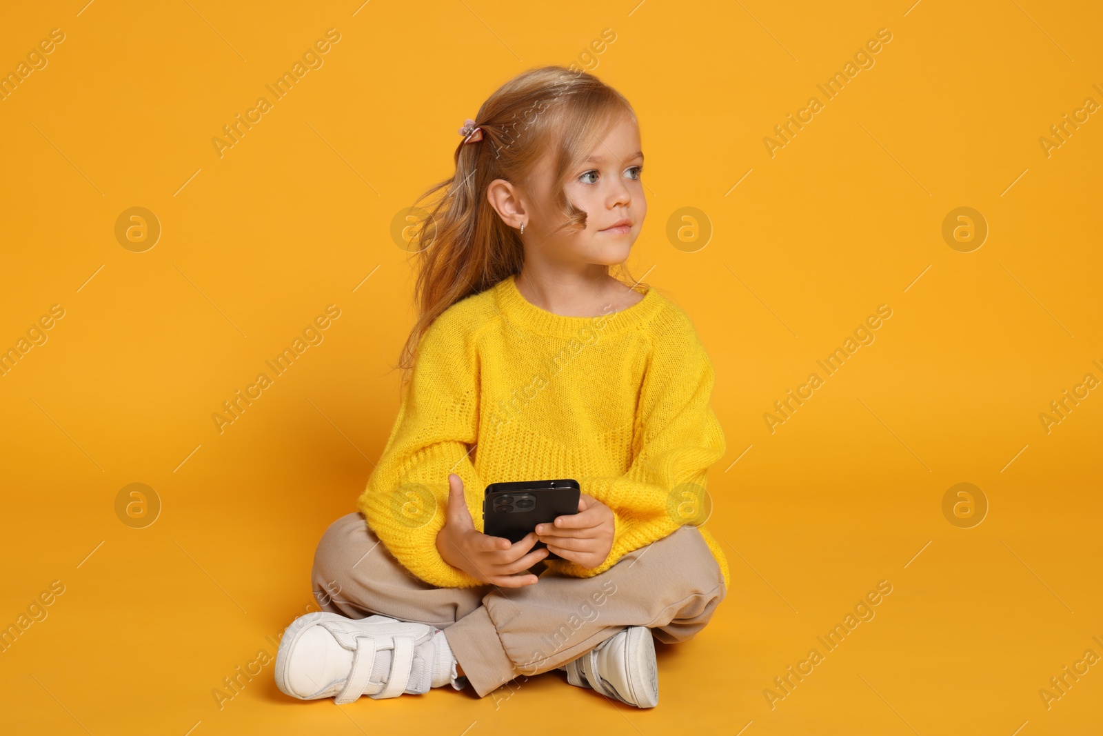 Photo of Cute little girl with smartphone against orange background