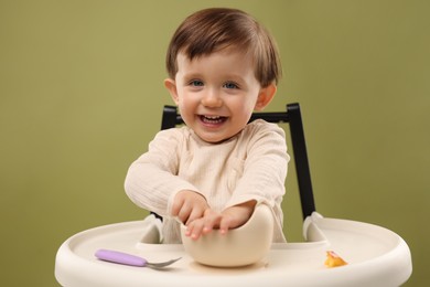 Photo of Cute little baby eating healthy food from bowl in high chair on olive background