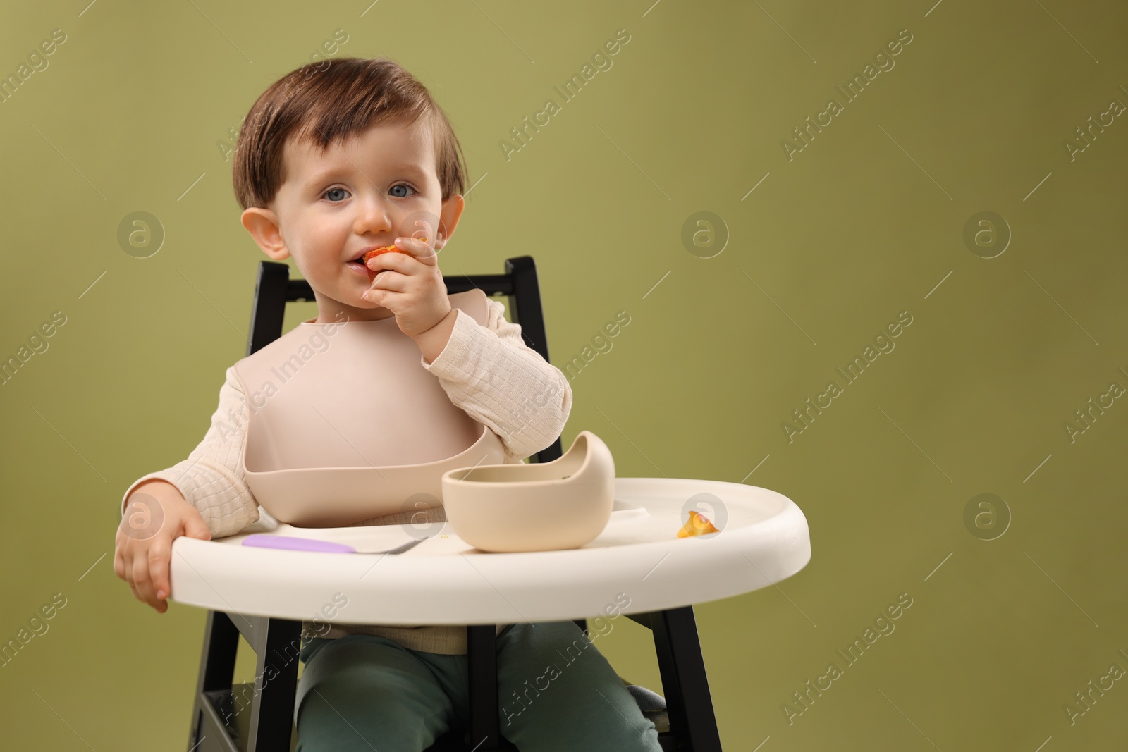 Photo of Cute little baby eating healthy food from bowl in high chair on olive background, space for text