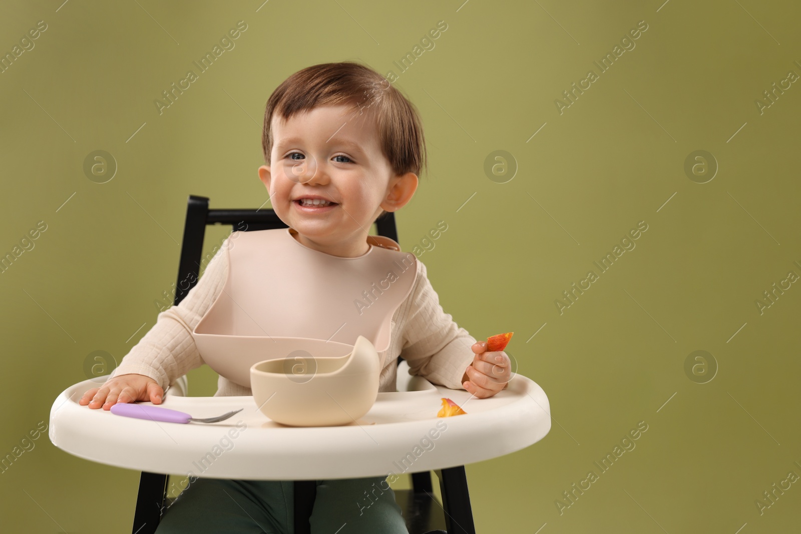 Photo of Cute little baby eating healthy food from bowl in high chair on olive background, space for text