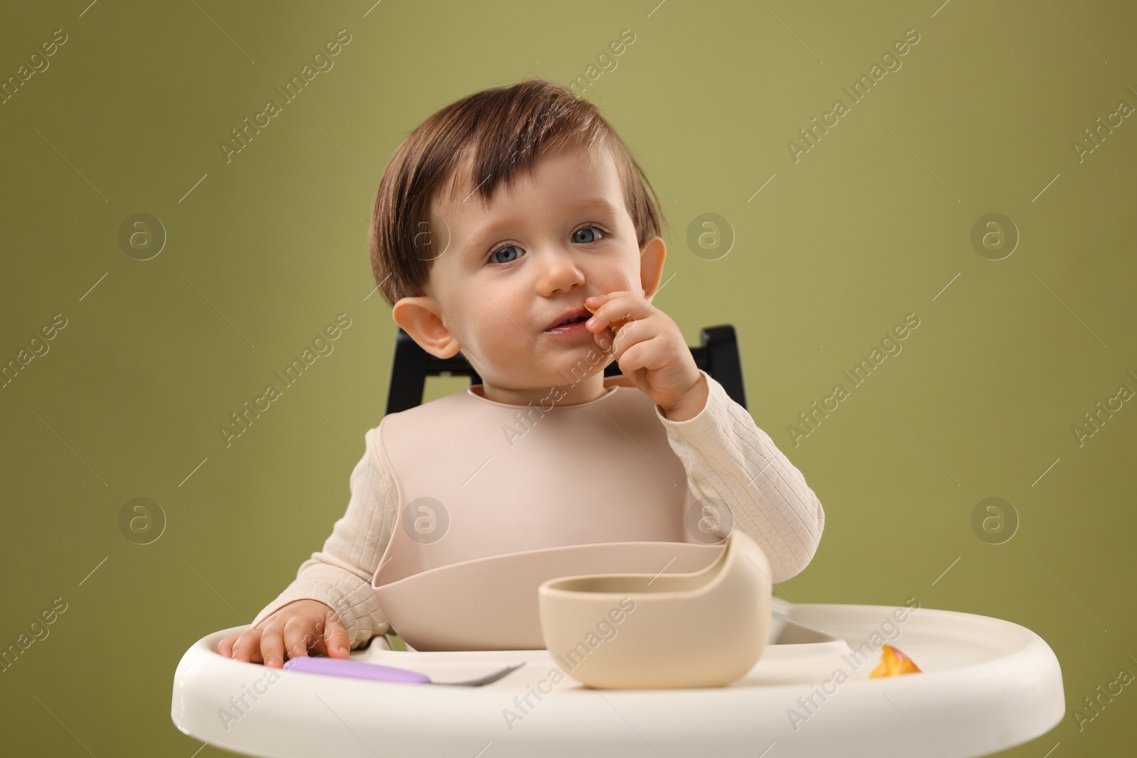 Photo of Cute little baby eating healthy food from bowl in high chair on olive background