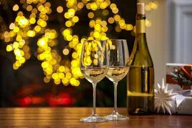 Photo of Glasses and bottle of white wine on wooden table against blurred Christmas lights. Bokeh effect