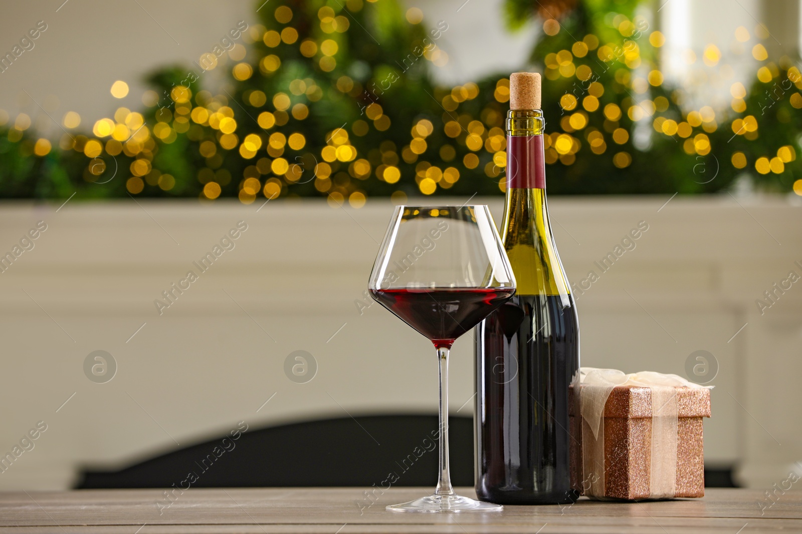 Photo of Red wine and gift box on wooden table against blurred Christmas lights, bokeh effect. Space for text