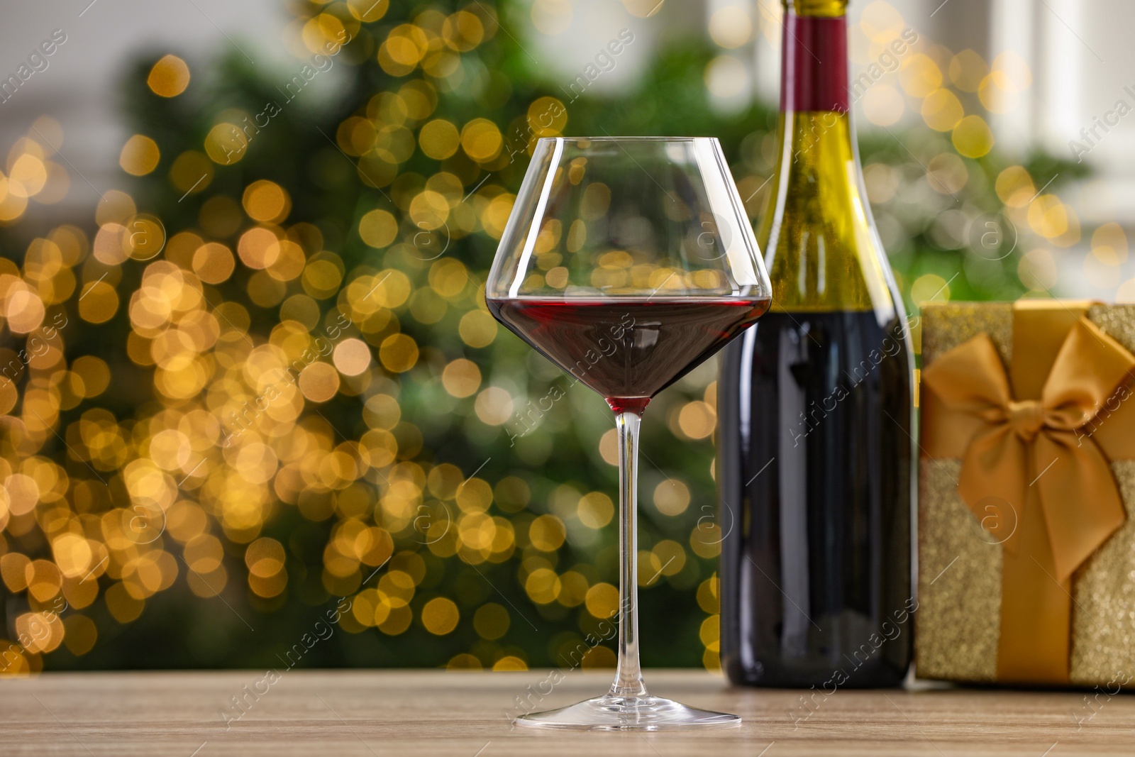 Photo of Red wine and gift box on wooden table against blurred Christmas lights, bokeh effect. Space for text