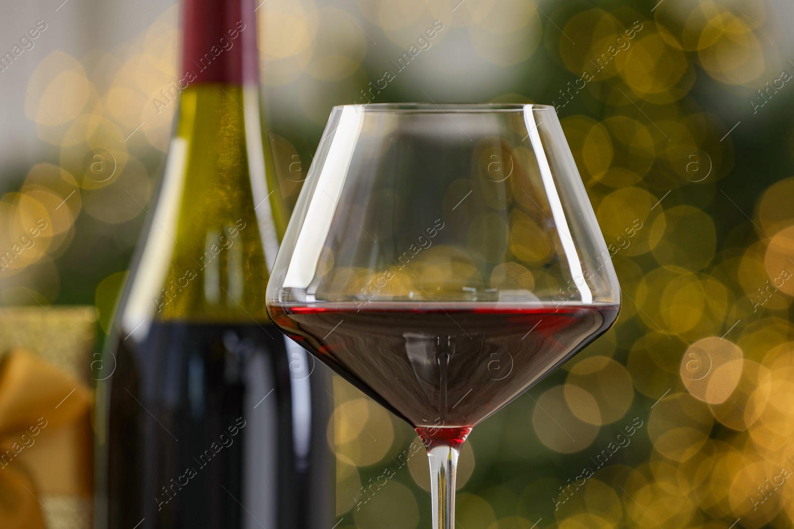 Photo of Glass and bottle of red wine against blurred Christmas lights, closeup. Bokeh effect