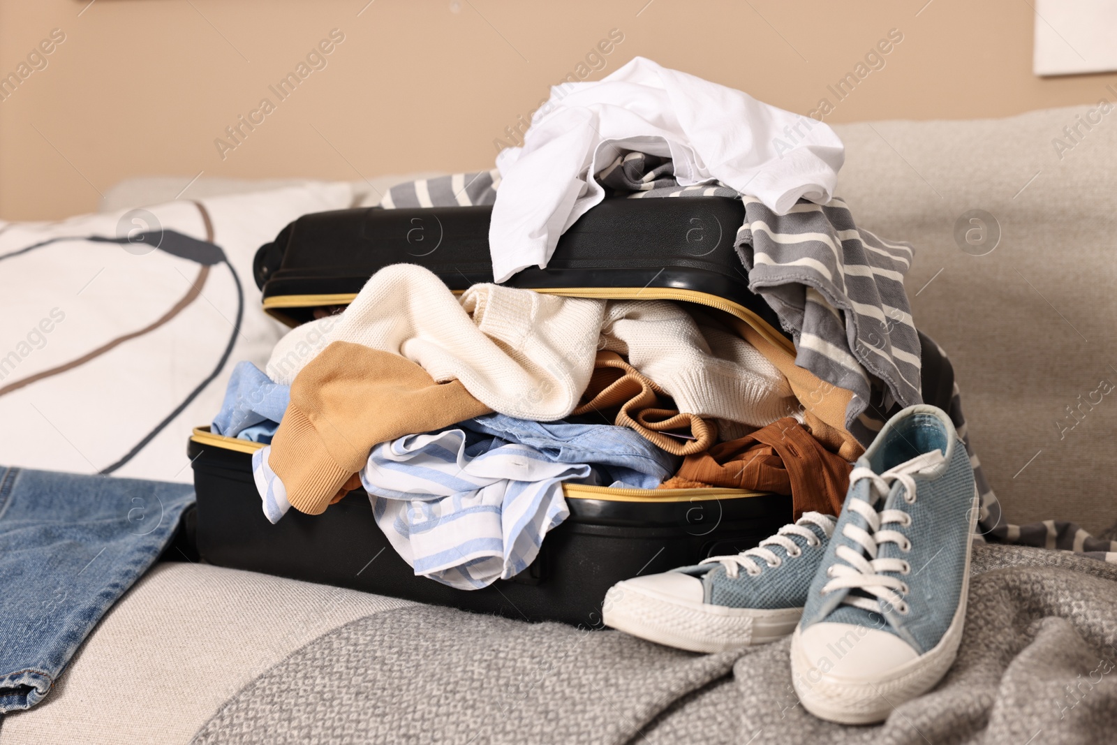 Open suitcase full of different messy clothes and sneakers on sofa indoors Photo of Open suitcase full of different messy clothes and sneakers on sofa indoors