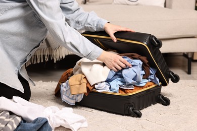 Photo of Woman trying to close suitcase full of different messy clothes indoors, closeup