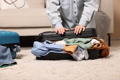 Photo of Woman trying to close suitcase full of different messy clothes indoors, closeup