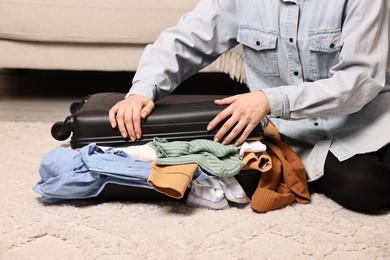 Photo of Woman trying to close suitcase full of different messy clothes indoors, closeup