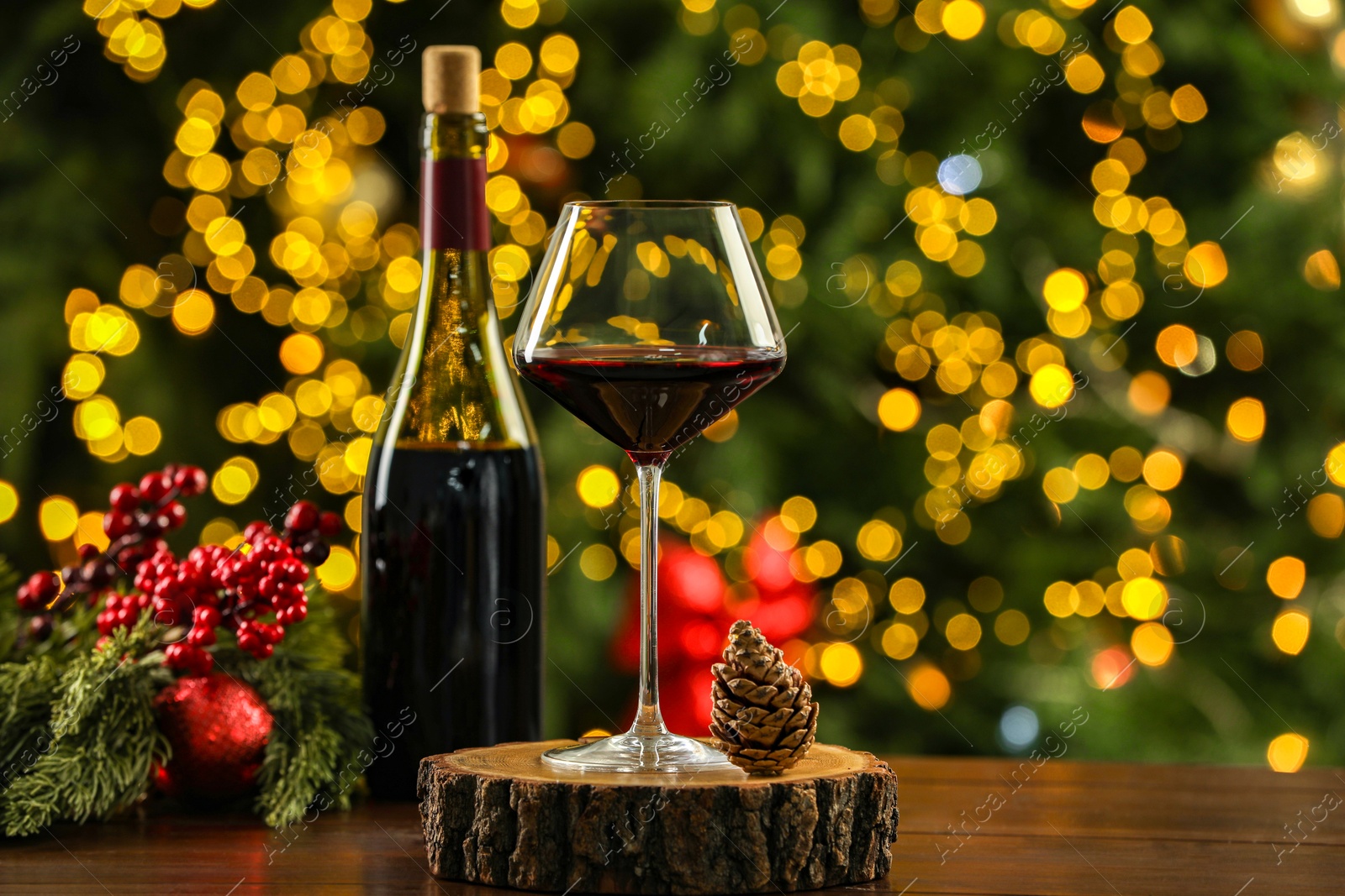 Photo of Glass and bottle of red wine with Christmas decor on wooden table against blurred lights. Bokeh effect