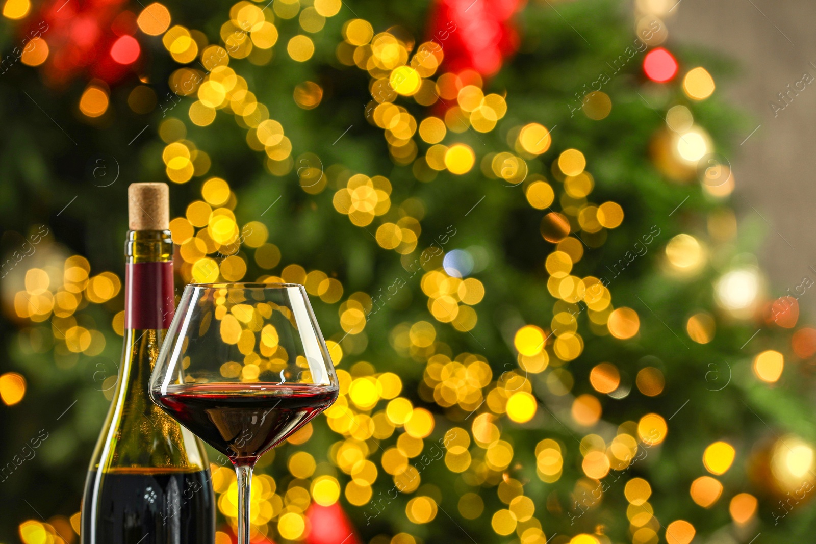 Photo of Bottle and glass of red wine against blurred Christmas lights, space for text. Bokeh effect