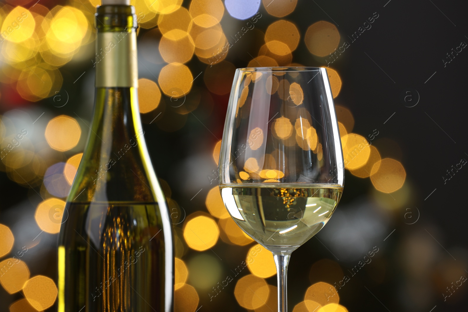 Photo of White wine in glass and bottle against blurred Christmas lights, closeup