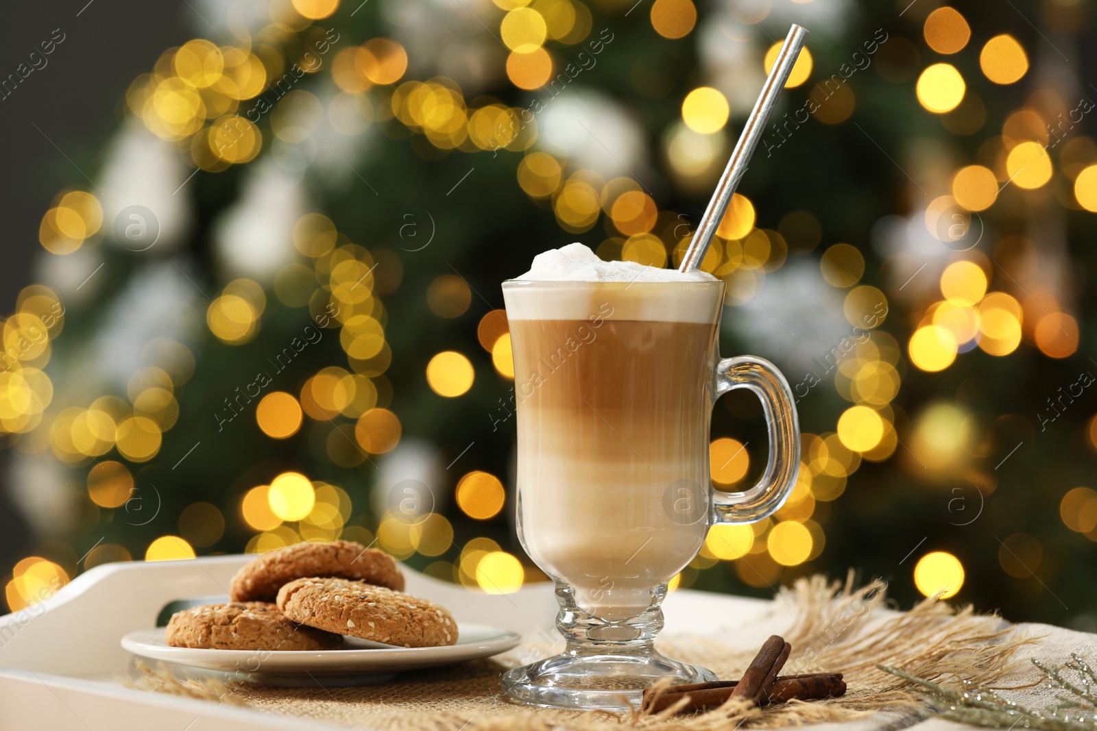 Photo of Aromatic coffee in glass, tasty cookies and cinnamon sticks on tray against blurred Christmas lights, bokeh effect