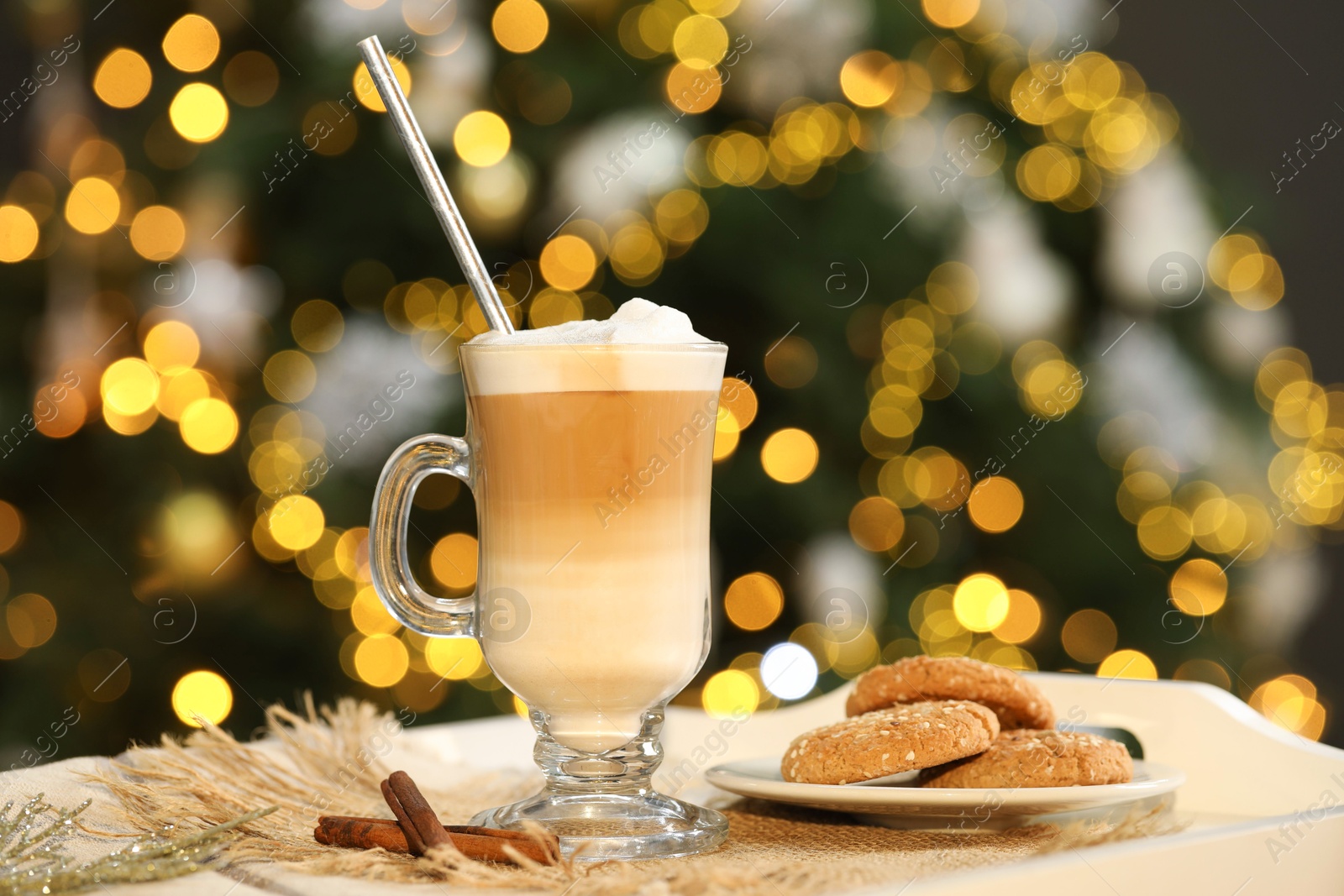 Photo of Aromatic coffee in glass, tasty cookies and cinnamon sticks on tray against blurred Christmas lights, bokeh effect