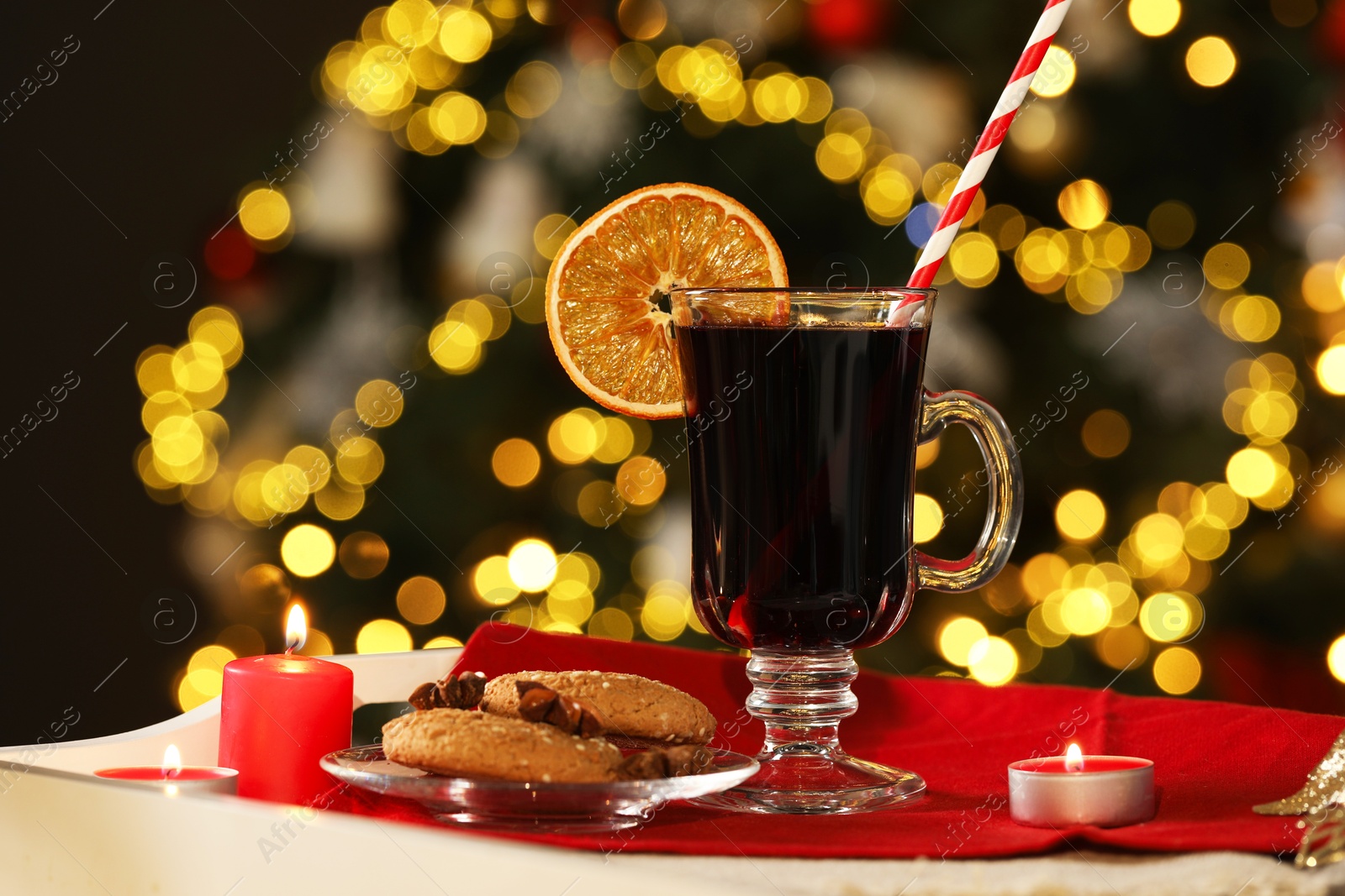 Photo of Aromatic mulled wine in glass cup, cookies and burning candles on tray against blurred Christmas lights, bokeh effect