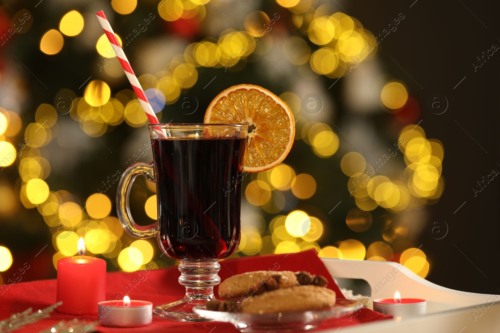 Photo of Aromatic mulled wine in glass cup, cookies and burning candles on tray against blurred Christmas lights, bokeh effect