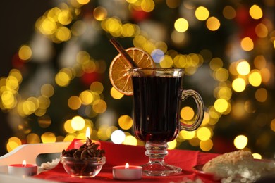 Photo of Aromatic mulled wine in glass cup, anise, cookies and burning candles on tray against blurred Christmas lights, bokeh effect