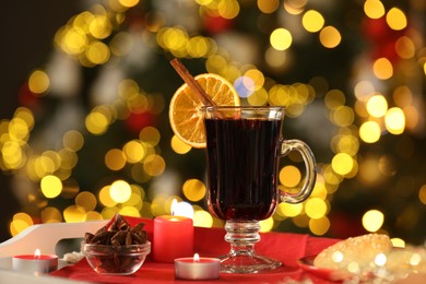 Photo of Aromatic mulled wine in glass cup, anise, cookies and burning candles on tray against blurred Christmas lights, bokeh effect