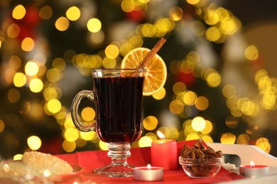 Photo of Aromatic mulled wine in glass cup, anise, cookies and burning candles on tray against blurred Christmas lights, bokeh effect