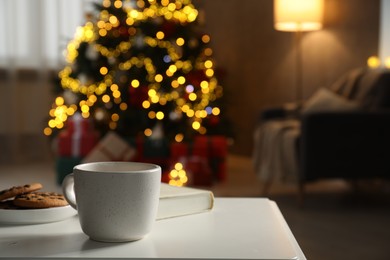 Photo of Cup of hot drink, book and cookies on white table in cozy room decorated for Christmas, closeup. Space for text