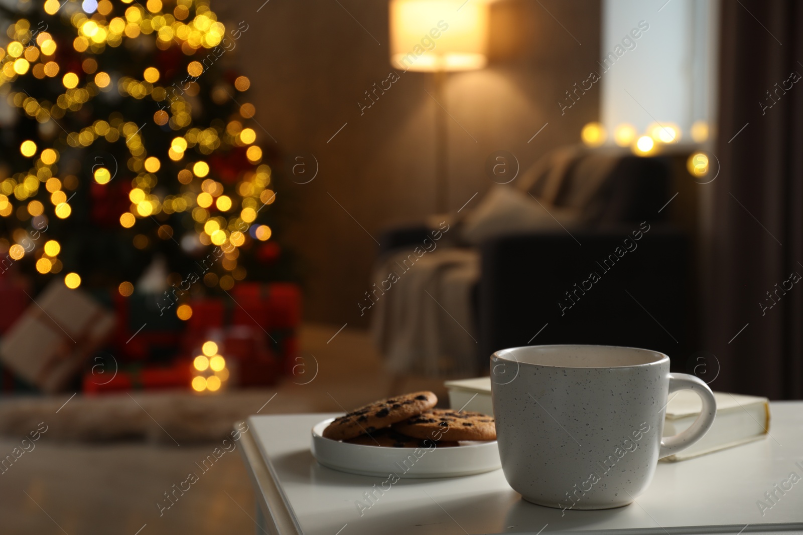 Photo of Cup of hot drink, book and cookies on white table in cozy room decorated for Christmas, closeup. Space for text