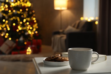 Photo of Cup of hot drink, book and cookies on white table in cozy room decorated for Christmas, closeup. Space for text