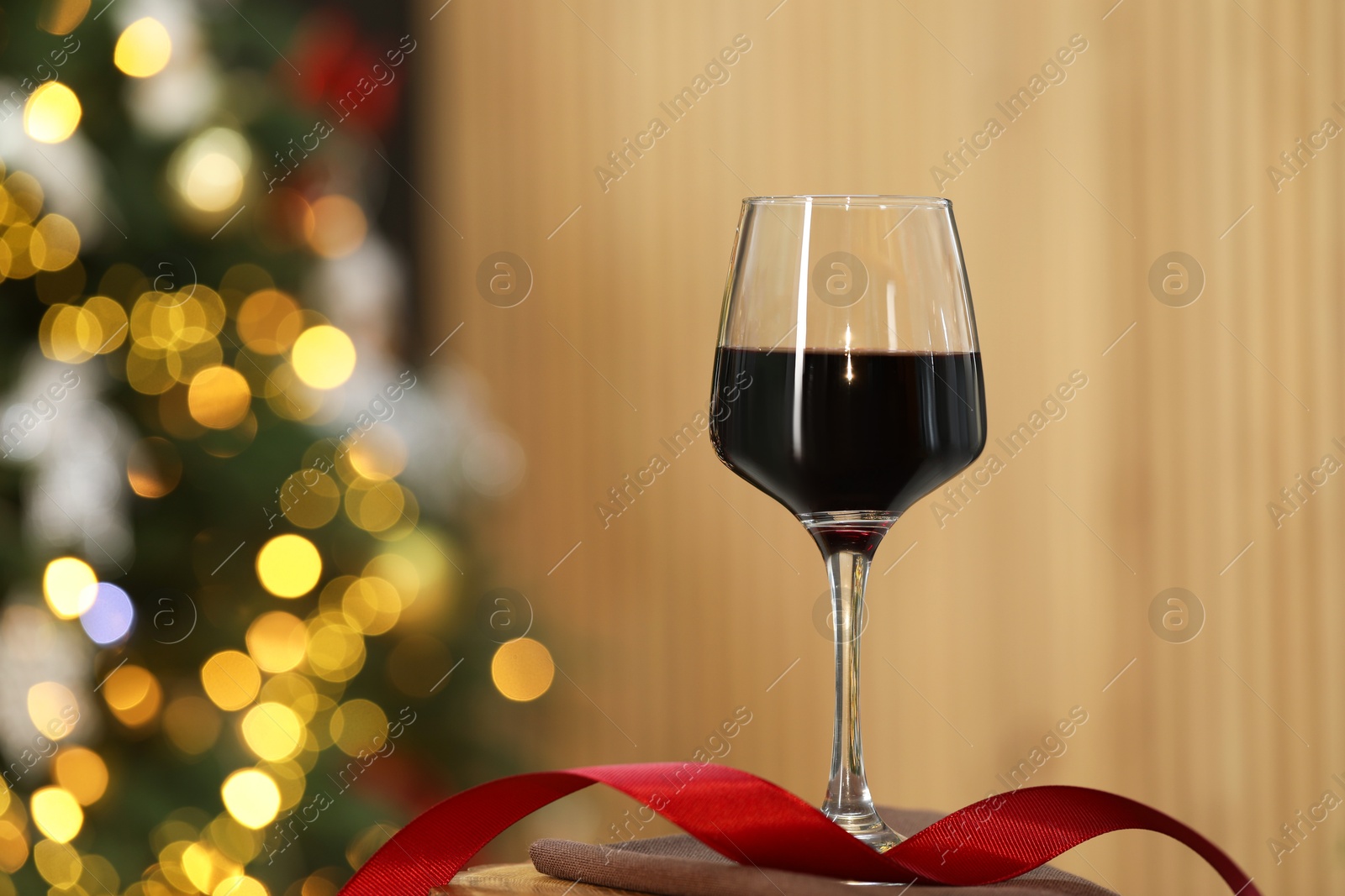 Photo of Red wine in glass and ribbon on table against blurred Christmas lights, space for text. Bokeh effect