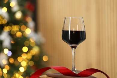 Photo of Red wine in glass and ribbon on table against blurred Christmas lights, space for text. Bokeh effect