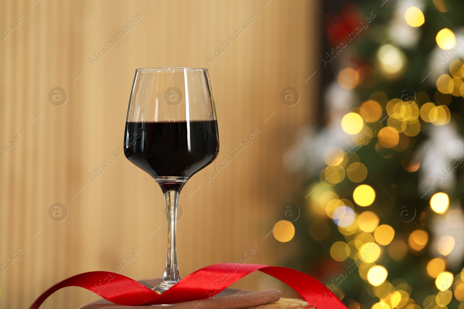 Photo of Red wine in glass and ribbon on table against blurred Christmas lights, space for text. Bokeh effect