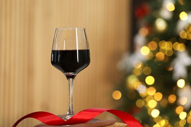 Photo of Red wine in glass and ribbon on table against blurred Christmas lights, space for text. Bokeh effect