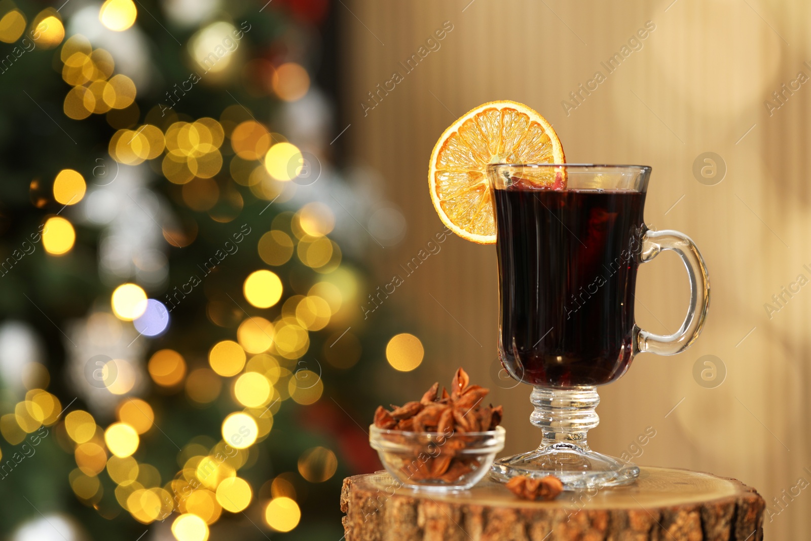 Photo of Aromatic mulled wine in glass cup and anise stars on stump against blurred Christmas lights, space for text. Bokeh effect