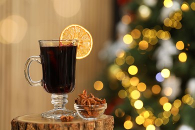 Photo of Aromatic mulled wine in glass cup and anise stars on stump against blurred Christmas lights, space for text. Bokeh effect