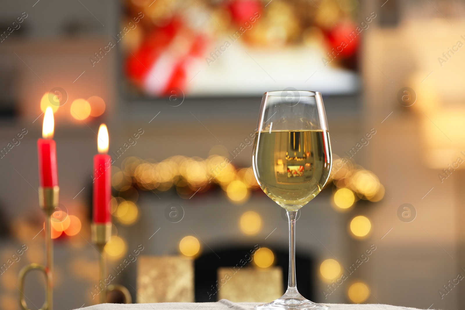 Photo of White wine in glass and burning candles on table against blurred Christmas lights, space for text. Bokeh effect