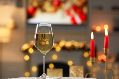 Photo of White wine in glass and burning candles on table against blurred Christmas lights, space for text. Bokeh effect