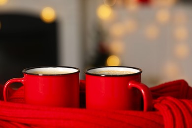 Photo of Tasty cocoa in cups and red scarf against blurred background with lights, closeup. Christmas drink