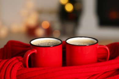 Photo of Tasty cocoa in cups and red scarf against blurred background with lights, closeup. Christmas drink