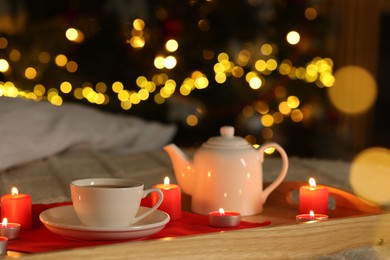 Photo of Aromatic tea in cup, teapot and burning candles on bed against blurred Christmas lights, closeup. Bokeh effect