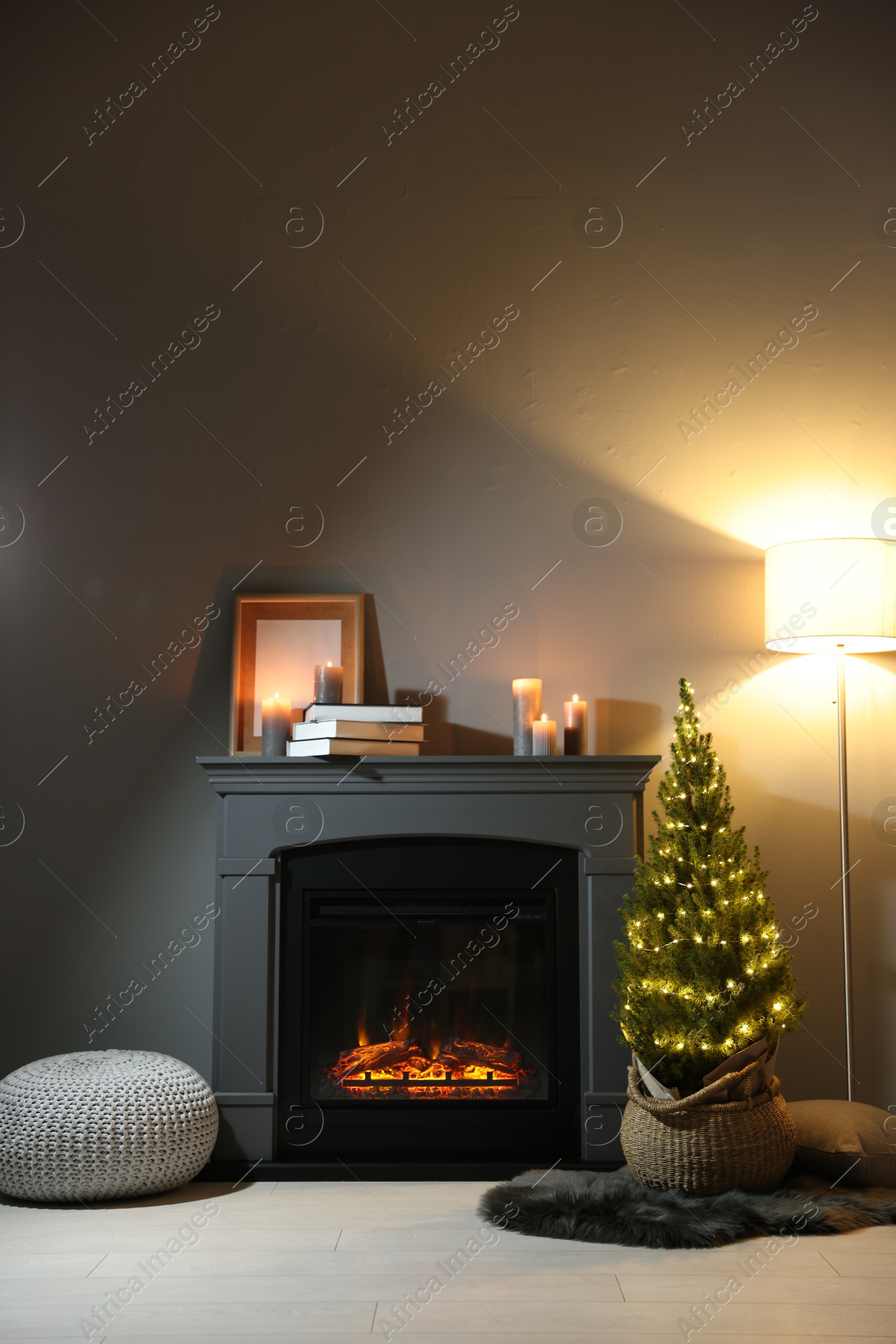 Photo of Small spruce tree in basket decorated with Christmas lights near fireplace indoors