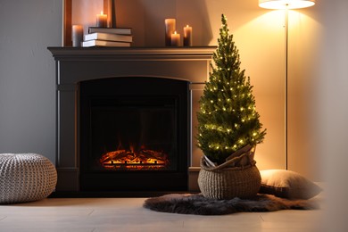 Photo of Small spruce tree in basket decorated with Christmas lights near fireplace indoors