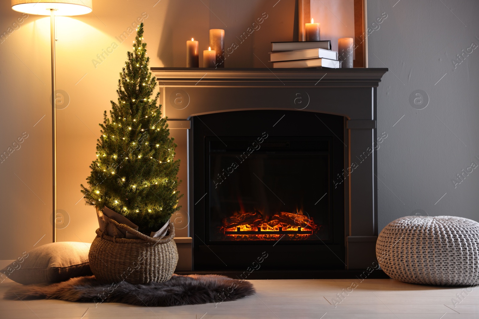 Photo of Small spruce tree in basket decorated with Christmas lights near fireplace indoors