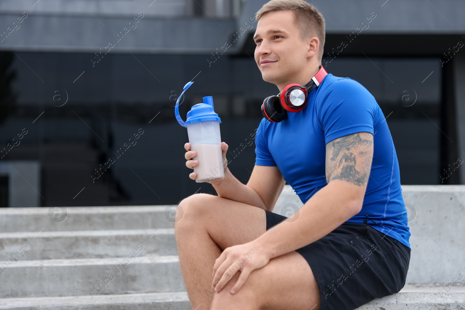 Photo of Athletic man with shaker of protein drink sitting on stairs outdoors. Space for text