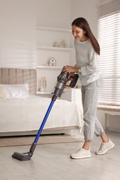 Photo of Young woman cleaning floor with cordless vacuum cleaner in bedroom