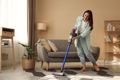 Photo of Smiling young woman cleaning rug with cordless vacuum cleaner in living room