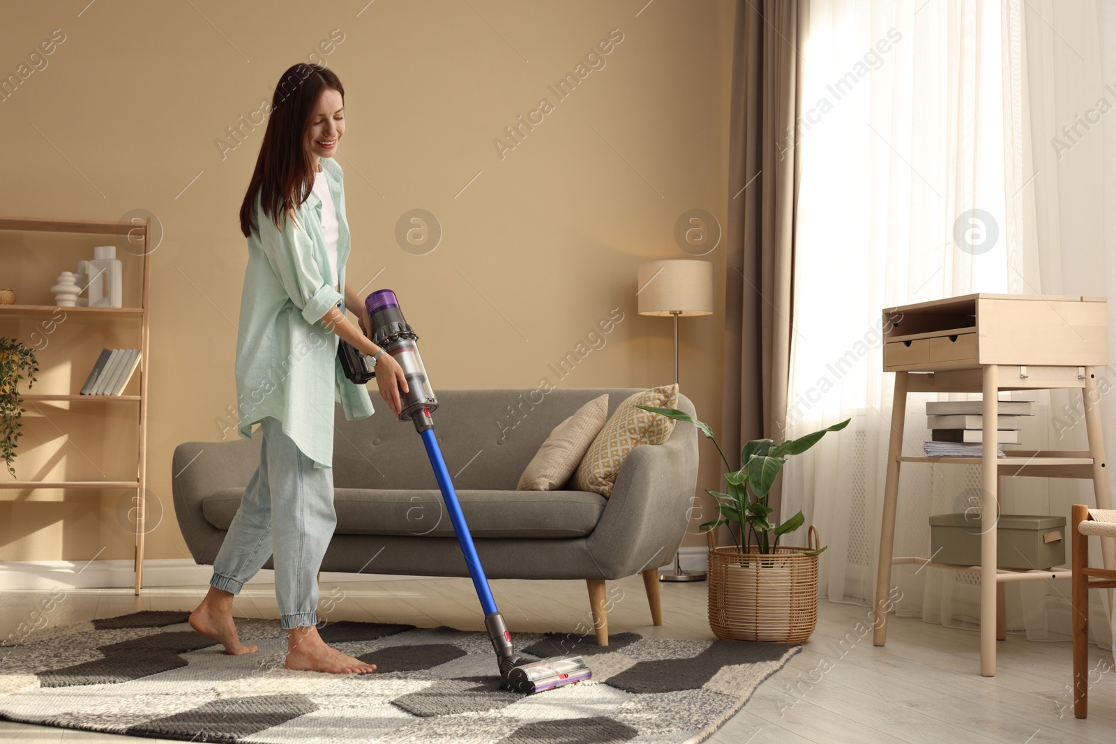 Photo of Smiling young woman cleaning rug with cordless vacuum cleaner in living room